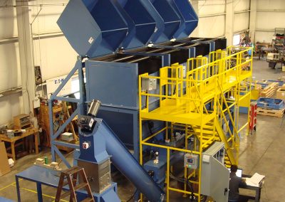 Four large container dumpers next to a custom extended bulk bag unloading frame with hoist and trolley, feeding four high-capacity live-bottom bins with horizontal agitators and a collection auger system with an integral drum-style metal separator, all controlled via a color touch screen.