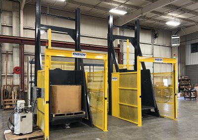Lift and Dump container dumper shown raising a Gaylord to an elevated discharge height, featuring an integrated weigh scale platform, secure perimeter guarding, and a light screen safety barrier; designed for high-capacity feeding of downstream equipment in recycling or processing facilities.