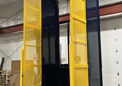 Tall bale dumper inside a recycling facility lifting a film bale to a 170-inch discharge height; compact 72-inch-wide frame with full perimeter guarding and a safety light screen, designed to feed bales directly into a shredder within a tight footprint.